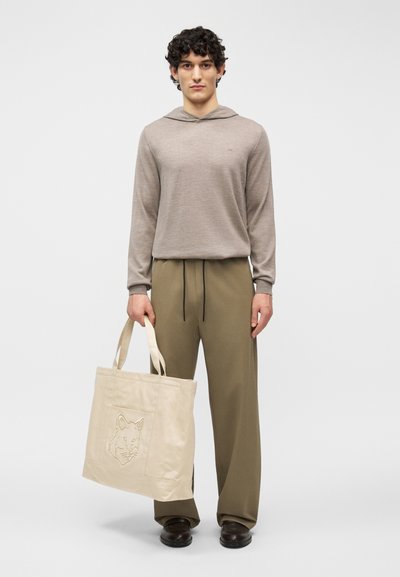 Νέος άνδρας με σγουρά μαλλιά, που φοράει μπεζ φούτερ με κουκούλα και χακί παντελόνι, κρατά μια μεγάλη κρεμ τσάντα tote με ανάγλυφο σχέδιο αλεπούς.