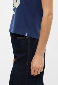 Ναυτικό μπλε βαμβακερό μπλουζάκι με άνετη εφαρμογή, με εκτυπωμένο σχέ διο στο μπροστινό μέρος και μικρή μπλε υφασμάτινη ετικέτα στο στρίφωμα. Συνδυασμένο με σκούρα τζιν παντελόνια.