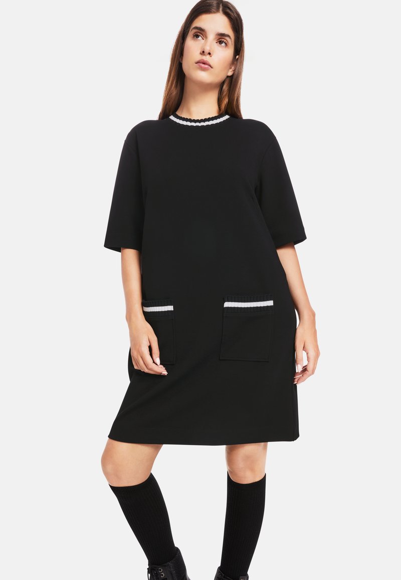 Schwarzes Kleid mit kurzen Ärmeln, runder Ausschnitt, verziert mit weißen gewellten Säumen und zwei Fronttaschen mit kontrastierenden gestreiften Details.