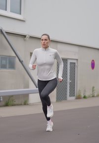 Haut de sport à manches longues gris clair avec une fermeture éclair quart, associé à un legging noir et des chaussures de course blanches, porté par une personne en mouvement.