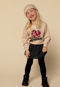 Lachend kind met een beige gebreide muts, een beige sweatshirt met een hart en de tekst "Dance", een zwarte gelaagde rok, zwarte panty's en zwarte laarzen.