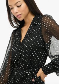 Czarna sukienka z przezroczystymi długimi rękawami, białe kropki, guziki z przodu i pasek do wiązania w talii. Miękki materiał o gładkiej fakturze.
