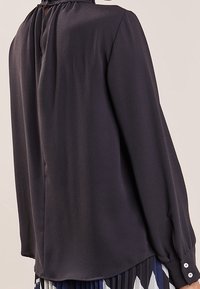Blusa nera a maniche lunghe con scollo arricciato e polsini con bottone, indossata con una gonna plissettata a motivi.