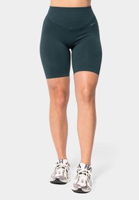 Hoog getailleerde, teal fietsbroek gemaakt van rekbare stof, met een gladde textuur en zonder zichtbare naden, gecombineerd met witte sportschoenen.
