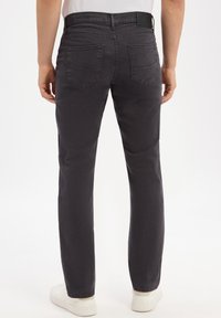 Uomo che indossa pantaloni dritti grigio scuro e sneaker bianche, visto da dietro su uno sfondo bianco puro.