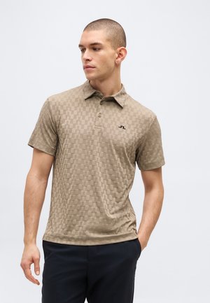 Mand iført beige kortærmet tekstureret poloshirt og mørke bukser, stående med venstre hånd i lommen, kigger mod venstre mod en ensfarvet baggrund.