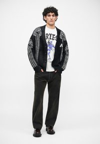 Cardigan negro con rayas grises y grandes botones, usado sobre una camiseta gráfica blanca. Combinado con pantalones oscuros y zapatos negros.