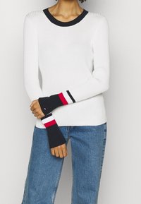 Person in einem weißen gerippten Pullover mit schwarzen, roten und weißen gestreiften Bündchen und Halsausschnitt, kombiniert mit blauen Jeans, vor einem schlichten Hintergrund stehend.