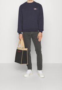 Marineblå sweatshirt med en liten grafik, grå jeans, vita sneakers och en beige och svart toteväska med röd text.