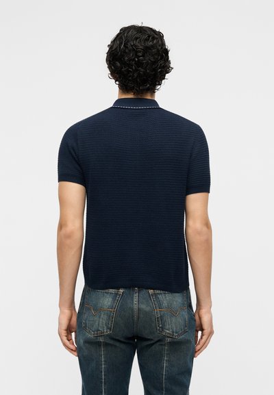 Άνδρας με σγουρά σκούρα μαλλιά που φοράει μπλε κοντομάνικο πόλο με υφή και ξεθωριασμένα μπλε τζιντζινάκια, στραμμένος πλάτη στην κάμερα.