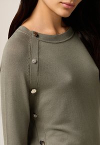Olivgrüner Pullover mit Rundhalsausschnitt, der dekorative silberne und kristallene Knöpfe entlang der Schulter für zusätzliche Detailverliebtheit aufweist.