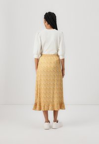 Gul blomstret midi-skjørt med flagrende kant, elastisk midjebånd, sammen med en hvit topp og hvite sneakers, fotografert fra en bakvinkel.