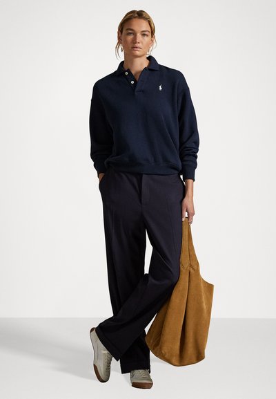 Pulover bleumarin cu guler și logo, asortat cu pantaloni negri și o geantă de mână din suedă bej. Pantofi sport cu accente bej și marron.