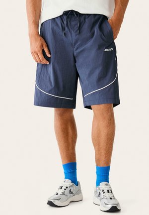 Muž nosi tmavomodré šortky s bielym lemovaním, modré ponožky a biele športové topánky, stojí so založenými rukami v vreckách.