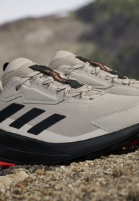 Jasnoszare buty sportowe z czarnymi akcentami i trzema ukośnymi paskami. Posiadają teksturowany materiał cholewki oraz solidną czarną podeszwę.