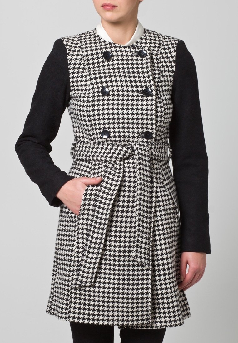 Mustavalkoinen houndstooth-takki pitkillä mustilla hihoilla, kaksirivinen malli ja vyötärövyö. Varustettu sivutaskulla ja suurilla napilla.