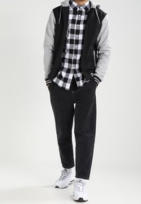 Grå och svart zip-up hoodie över en svartvit rutmönstrad skjorta, kombinerad med svarta byxor och vita sneakers med grå detaljer.