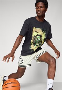 Melnā T-kreklā ar košu grafisku dizainu, uz kura attēlots profils, apvienots ar gaišām, perforētām basketbola šortiem un oranžu basketbola bumbu.