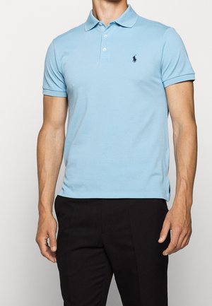 Homme portant un polo bleu clair à manches courtes avec un petit logo foncé sur la poitrine et un pantalon noir, debout devant un fond uni.