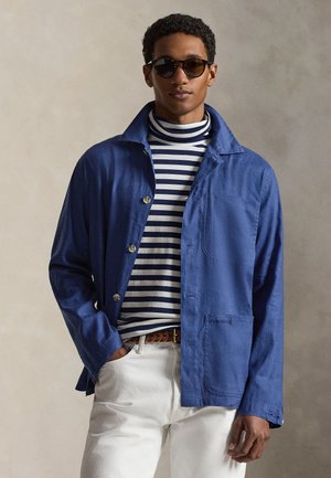 Άνδρας που φοράει σκοτεινά γυαλιά ηλίου, μπλε σακάκι με κουμπιά πάνω από μπλε και άσπρο ριγέ ζιβάγκο, άσπρο παντελόνι και καφέ πλεκτή ζώνη.