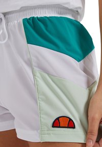 Pantalones cortos blancos con acentos en verde azulado y verde claro, presentando un diseño en bloque de colores. Logo bordado en naranja y negro en el dobladillo.