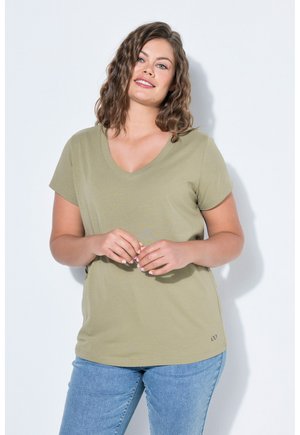 Femme souriante aux cheveux bruns ondulés portant un t-shirt vert olive à col en V et un jean bleu clair, debout contre un fond blanc.