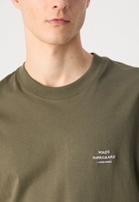 Olivovo zelené bavlnené tričko s klasickým okrúhlym výstrihom, s vyšitým textom "MADS NØRGAARD COPENHAGEN" v bielej farbe na ľavej strane hrudi.