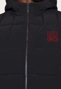 Schwarze gepolsterte Weste mit einem Reißverschluss, Kapuze und rotem "Hugo"-Logo. Glatte Textur mit gestepptem Muster über dem Stoff.