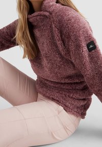 Pullover di pile viola morbido con colletto alto e patch in tessuto nero sulla manica, abbinato a pantaloni aderenti rosa chiaro.