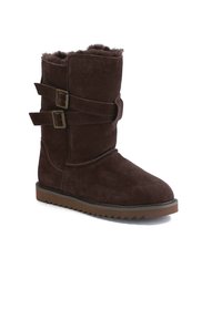 Botas de ante marrón hasta la mitad de la pantorrilla con punta redonda, que presentan dos correas con hebillas y una suela de goma texturizada para agarre y estabilidad.