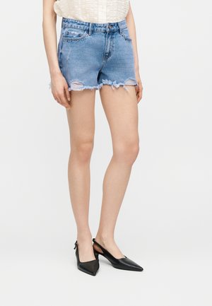 Kvinde iført lyseblå slidte denimshorts med frynset kant og sorte spidse slingback-hæle, stående mod en ensfarvet baggrund.