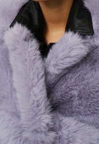 Persona con un cappotto di pelliccia sintetica fluffy lavanda sopra una giacca nera in pelle con colletto, con il colletto del cappotto ben visibile.