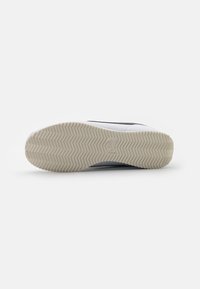 Fehér sneaker, bordázott gumitalppal, amely halas mintázattal rendelkezik, és fekete részletekkel az oldalakon. Simább felső anyag.