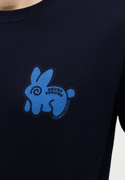 Γραφικό μπλε στυλιζαρισμένου κουνελιού σε μπλε σκούρο πουκάμισο με κείμενο "NEVER ASSUME" και "PS PAUL SMITH" κοντά στην ουρά του κουνελιού.