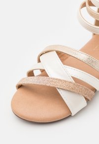 Sandaler med stropper som har hvitt lær og metalliske gullakenter. Sålen er beige med en myk tekstur, og stroppene er vevd sammen.