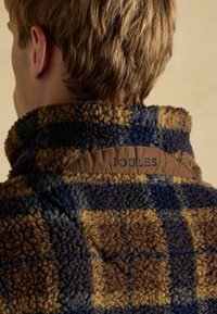 Plush, check-pattern fleece jacket in blue, brown, and tan tones. Features a high collar and a brown label embossed with "JOULES."