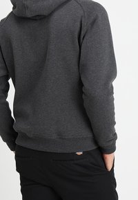 Sudadera gris oscuro con un bolsillo tipo canguro, puños acanalados y cintura ajustada. La tela parece suave y texturizada; presenta un logotipo en la parte posterior.