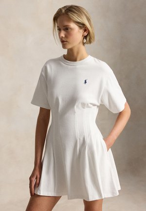 Femme en robe blanche à manches courtes avec plis et petit logo brodé bleu marine, debout avec une main dans la poche, fond neutre.