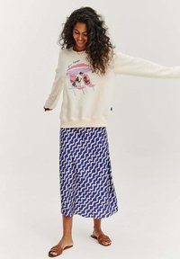 Bílá mikina se grafickým potiskem psů na plážových lehátkách, která je zkombinována s dlouhou modrou sukní s bílým vzorem vln. Hnědé sandály.