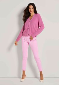 Vrouw in een roze cardigan met knopen, lichtroze broek en witte hoge muiltjes poseert tegen een lichtgrijze muur.