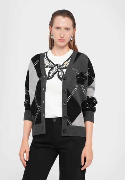 Grå cardigan med svart og grå diamantmønster, ribbede mansjetter og knapper. Brukt over en hvit skjorte med et svart og gult skjerfprint.