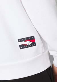 Vit sweatshirt med en Tommy Hilfiger-logotyp som visar två händer som skakar hand, en röd och en vit, med text ovanför och nedanför händerna.