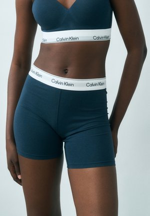 Γυναίκα που φοράει σκούρο μπλε bralette Calvin Klein και αντίστοιχα boxer σορτς με λευκά ελαστικά κορδόνια με το λογότυπο.