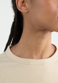 Beige t-shirt with a round neckline, made of soft fabric, showing the collar and neck area. Slight texture visible on the fabric.