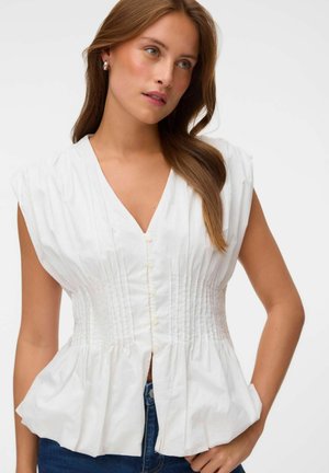 Jeune femme portant un chemisier blanc sans manches boutonné avec une taille froncée et un jean en denim, posant avec une main dans la poche.
