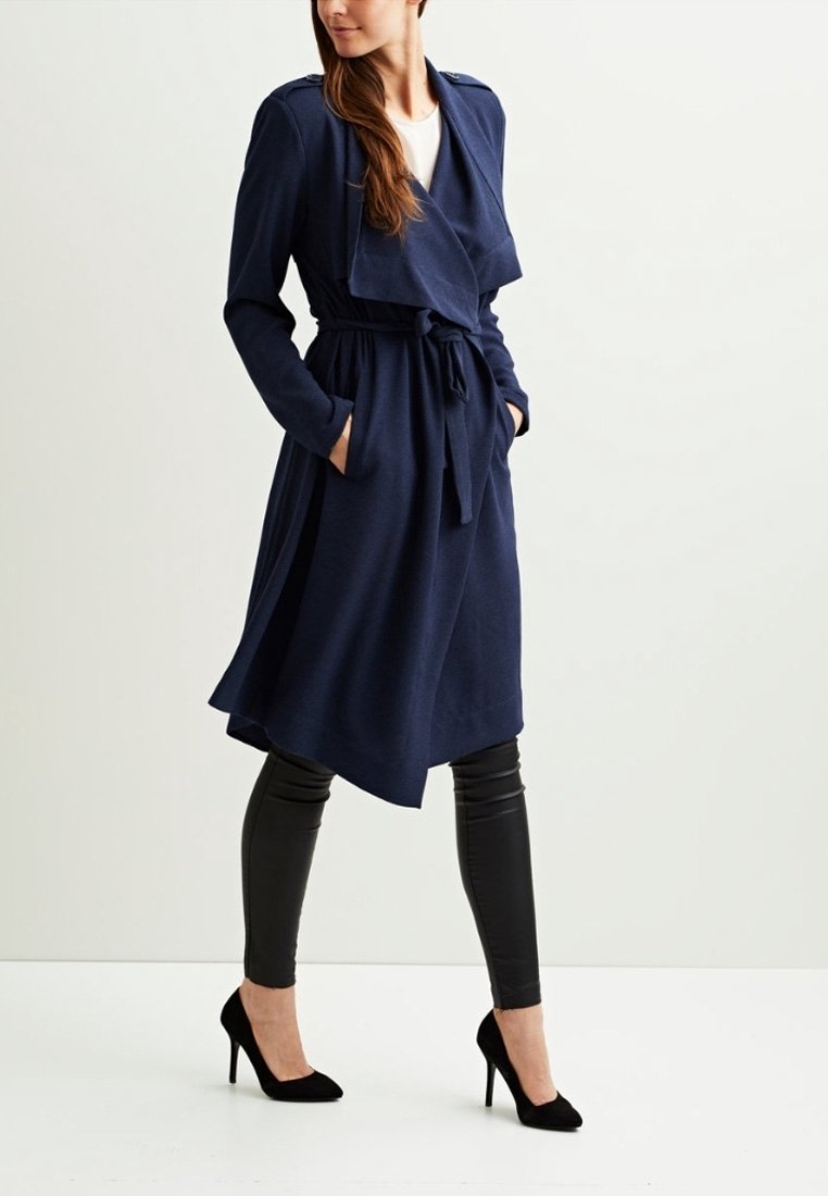 Femme portant un trench-coat bleu marine ceinturé sur des leggings noirs et des talons hauts noirs, debout les mains dans les poches.
