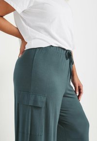 Lösa turkosa byxor med resårmidja och snörning. Har en sidoficka och en mjuk, slät textur. Matchas med en vit t-shirt.