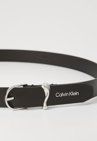 Juodas odinis diržas su sidabriniu metaliniu sagtimi ir iškilniu "Calvin Klein" logotipu ant stačiakampio ženklelio. Lygus paviršius, minimalistinis dizainas.