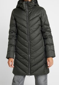 Mörkgrön, vadderad puffjacka med chevronmönster, hel dragkedja, huva och långa ärmar. Slät, vattenavvisande tyg.