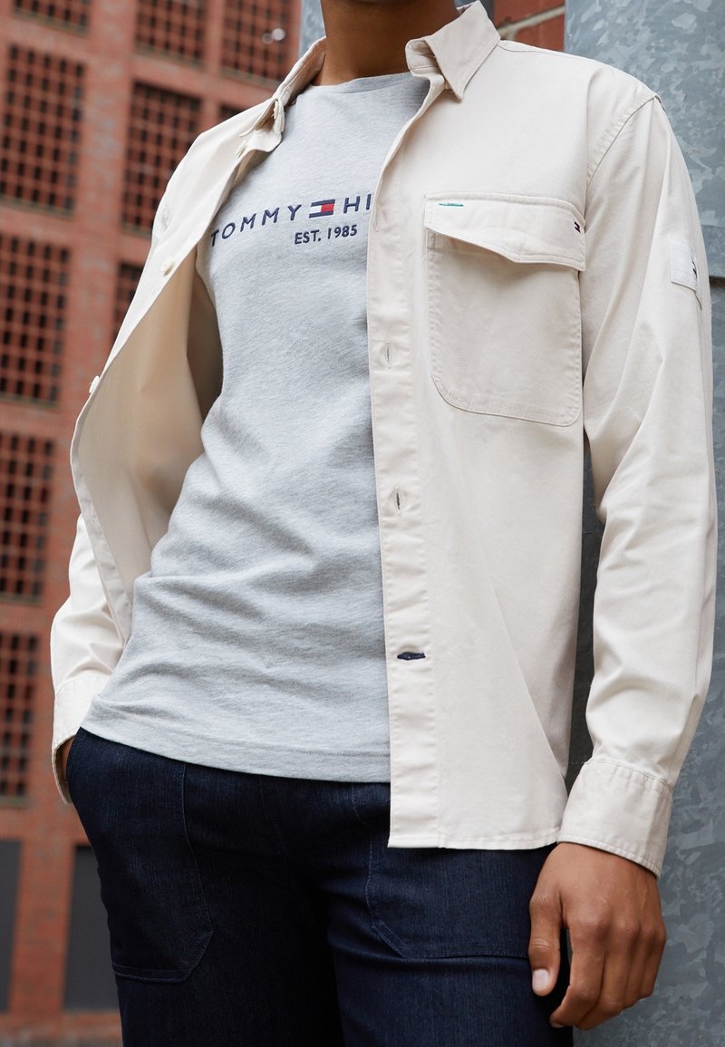 Beige skjorte med knaplukning og en enkelt brystlomme over en grå t-shirt med blå og rød branding, parret med mørke jeans.